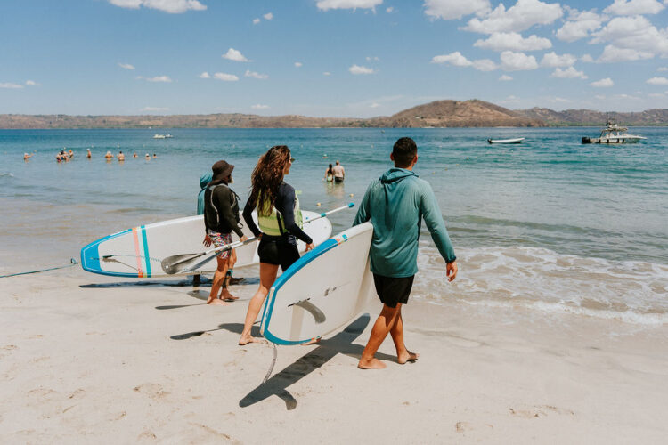 Three people carrying paddleboards walking in the sand toward the ocean