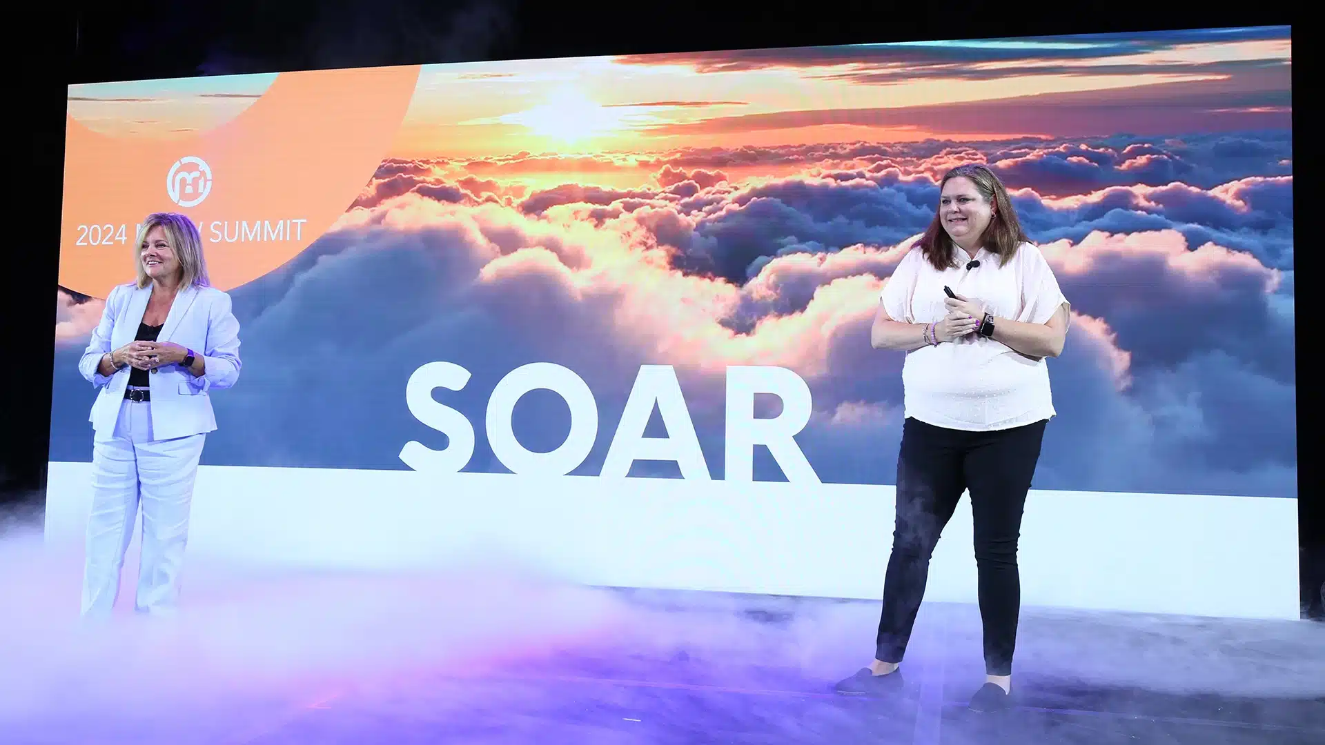 Two women on stage with the backdrop of clouds and the word "Soar"