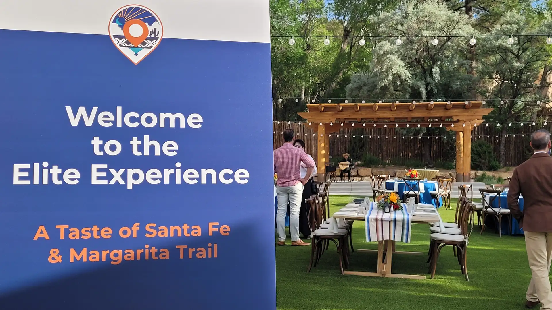 Elite experience signage in foreground with the outdoor event in background with beautiful table scape, pergola with hanging lights and a musican.