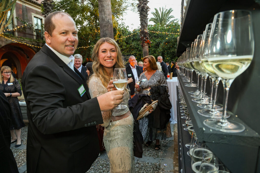 Two attendees standing next to a champagne wall smiling and raising their glasses of champagne